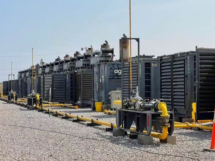 Row of skid-mounted natural-gas generator sets lined up along a yellow gas pipeline manifold on a gravel pad at the Alberta site.