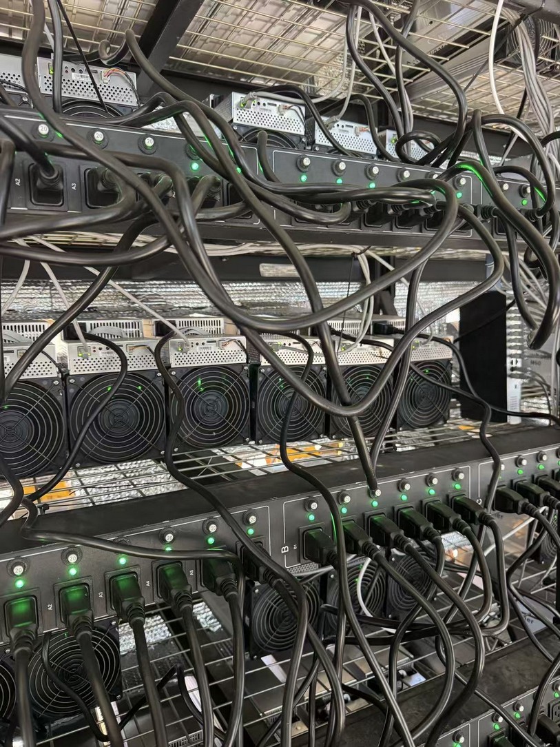 Interior of the compute hall showing racked mining machines with green status lights and cabled power distribution units.
